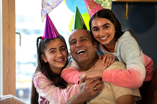 A father and his two daughters wearing casual clothing in a holiday rental accommodation located in Amble, Northumberland. They are gathered to celebrate the teenage girl's birthday and are wearing party hats in a room filled with balloons and bunting. They look and smile at the camera.