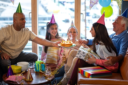 A multiracial, multi-generational family wearing casual clothing in a holiday rental accommodation located in Amble, Northumberland. They are gathered to celebrate the teenage girl's birthday and are wearing party hats in a room filled with balloons and bunting. They all watch and smile as the girl blows out the candles on her birthday cake.