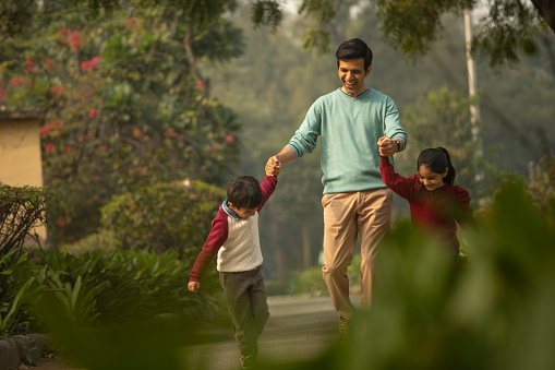 Cheerful playful children holding and pulling young smiling father's hands while walking in park during weekend
