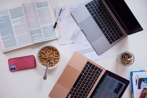 Directly above view of the desk of a university student, on the desk is two laptops, surrounding the laptops is a bowl of cereal, textbooks, paperwork, pencils and a smartphone.