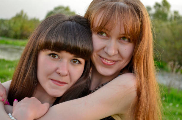Two sisters enjoy a playful interaction, standing closely together with one smiling brightly while the other gazes affectionately. The cheerful atmosphere is enhanced by a vivid pink backdrop.