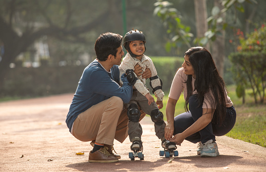 Mother and father holding son's hands and assisting him in roller skating on footpath against trees in park during weekend
