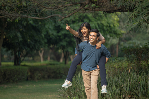 Happy young husband piggybacking cheerful laughing wife wile walking against trees in park during weekend