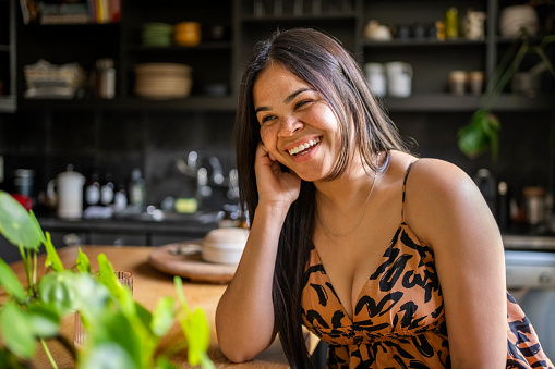 Portrait of pretty young woman looking away and smiling while sitting at table in kitchen at home