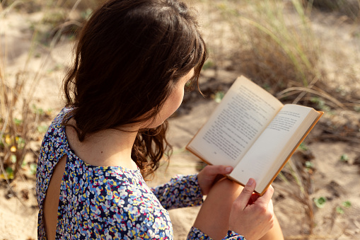 Young woman wearing a blue dress, sitting in the sand at the beach dunes reading a yellow hard cover book