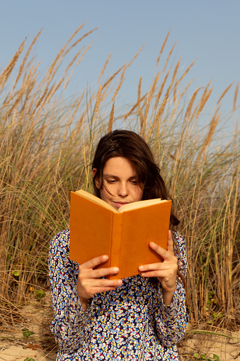 Young woman wearing a blue dress, sitting in the sand at the beach dunes reading a yellow hard cover book