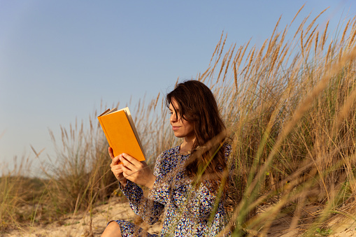 Young woman wearing a blue dress, sitting in the sand at the beach dunes reading a yellow hard cover book