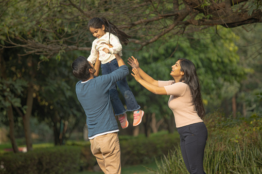 Cheerful young mother with arms raised looking at father throwing daughter in air while enjoying weekend against trees in park