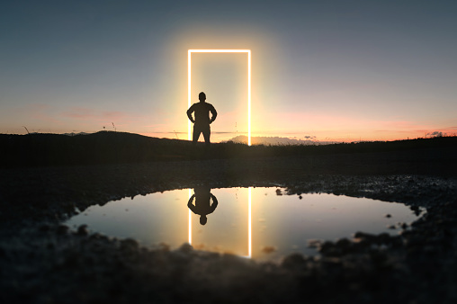 A glowing yellow neon gate in a natural area at dusk, with a man's silhouette facing it. Reflection on a puddle.