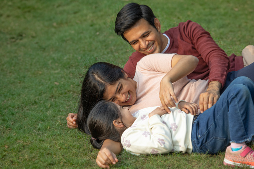 Playful young parents tickling cheerful daughter while lying on grassy field in park during weekend