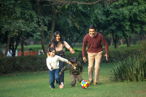 Cheerful young parents running behind children playing with ball on grassy field in park during weekend