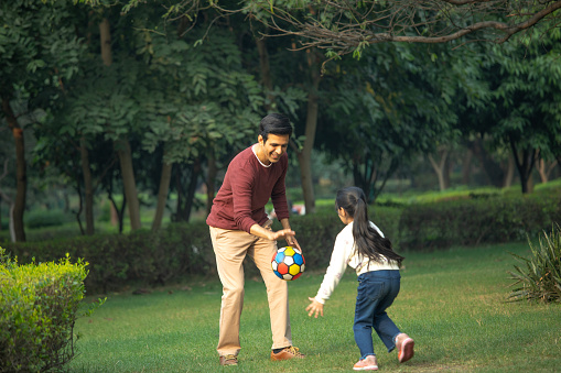 Cheerful father and daughter playing with ball on grassy field against trees in park during weekend