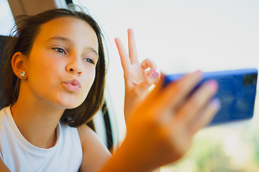 Little smiling girl travels by train while communicating via video conference on phone, takes selfie photo with air kiss.
