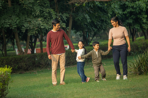 Young mother and father holding happy children's hands and walking together while spending weekend in park