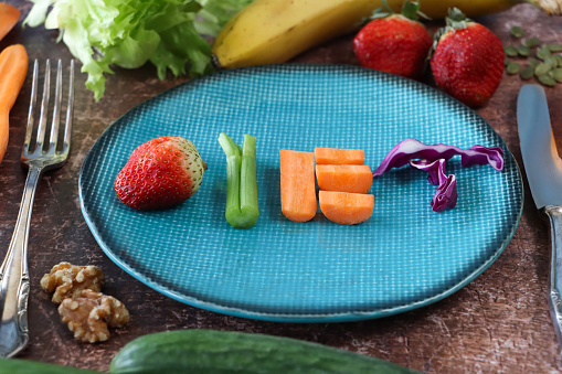 Stock photo showing close-up, elevated of a healthy eating and diet concept depicted by a plate containing fruit and vegetables used to form the letters of the word 'diet'. The letter 'D' is made using a strawberry, a celery stick and almond nut form the 'I', a chopped carrot creates an 'E' whilst shreds of red cabbage are the 'T'. Surrounding the plate on a brown background is a display of walnut halves, banana, cucumbers, strawberries, lettuce, carrot and pumpkin seeds.