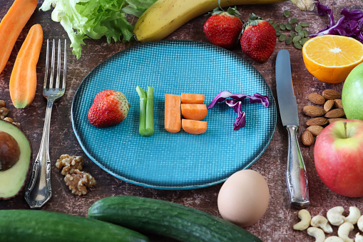 Stock photo showing close-up, elevated of a healthy eating and diet concept depicted by a plate containing fruit and vegetables used to form the letters of the word 'diet'. The letter 'D' is made using a strawberry, a celery stick and almond nut form the 'I', a chopped carrot creates an 'E' whilst shreds of red cabbage are the 'T'. Surrounding the plate on a brown background is a display of cashew nuts, walnut halves, banana, red and green apples, egg, cucumbers, avocado half, half an orange, strawberries, lettuce, carrot and red cabbage.