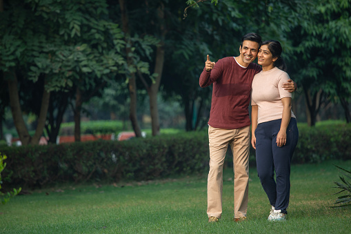 Young smiling husband pointing and looking away with wife while spending leisure time in park during morning