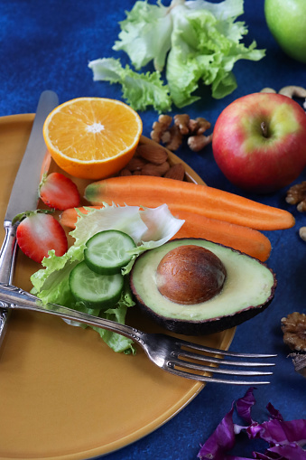 Stock photo showing an intermittent fasting concept depicted by a plate and cutlery forming a clock face and hands with the smallest section on the plate being filled with a pile of almonds, half an orange, strawberry cut in two, a sliced carrot, lettuce leaves, slices of cucumber and half an avocado displaying it's large seed. Surrounding the plate on a blue background is a display of cashew nuts, walnut halves, banana, red and green apples, egg, potatoes and red cabbage.