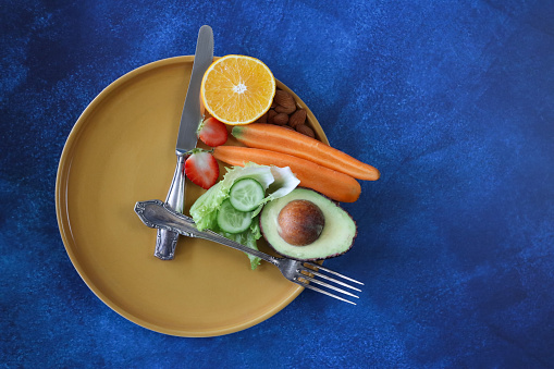 Stock photo showing an intermittent fasting concept depicted by a plate and cutlery forming a clock face and hands with the smallest section on the plate being filled with a pile of almonds, half an orange, strawberry cut in two, a sliced carrot, lettuce leaves, slices of cucumber and half an avocado displaying it's large seed.