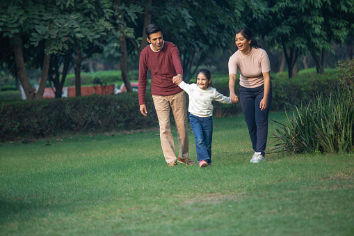 Smiling playful daughter holding and pulling parents hands while walking on grassy field in park during weekend