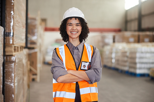 Portrait of smiling young female engineer wearing a hardhat and reflective vest standing with her arms crossed in a large shipping and distribution warehouse