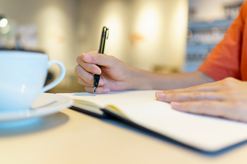 Close-up of an Asian woman seated at a table, focused on her work. She is holding pens and a personal organizer, diligently making notes in documents spread out in front of her. In the midst of her tasks, she enjoys a cup of coffee, creating a balanced and productive workspace