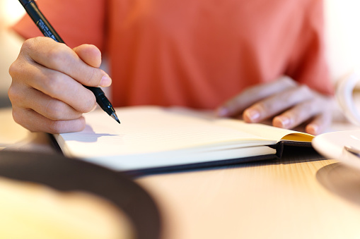 Close-up of an Asian woman seated at a table, focused on her work. She is holding pens and a personal organizer, diligently making notes in documents spread out in front of her. In the midst of her tasks, she enjoys a cup of coffee, creating a balanced and productive workspace