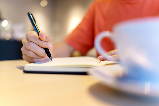 Close-up of an Asian woman seated at a table, focused on her work. She is holding pens and a personal organizer, diligently making notes in documents spread out in front of her. In the midst of her tasks, she enjoys a cup of coffee, creating a balanced and productive workspace