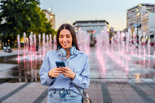 Happy smiling woman enjoying while using phone in the city with fountain in the background
