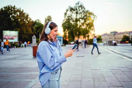 Happy smiling woman with headphones enjoying while using phone in the city