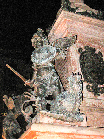 Statue of a small angel fighting a cockatrice, representing Pestilence, at the base of the Mariensäule, in Munich Germany, at night. The Mariensäule in Munich was erected in 1638 to celebrate the end of the Swedish occupation of the city during the Thirty Years War. The pedestal is surrounded by statues of four putti, each fighting a beast, which symbalise a n adversity the city had to othercome,