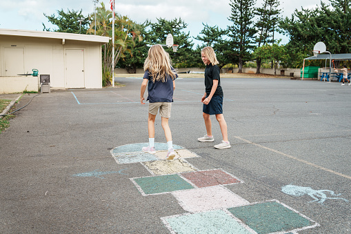 Two young school girls wearing school uniforms play hopscotch with one another outside during a class recess.