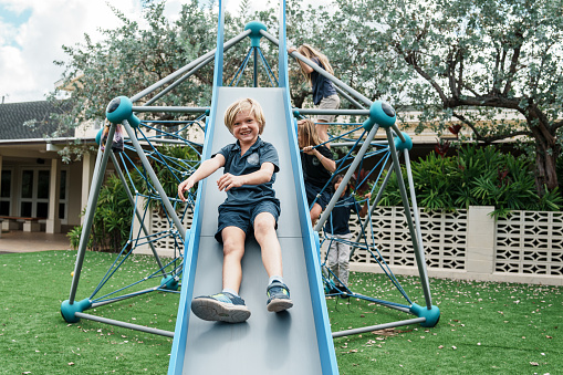 A happy young schoolboy of Caucasian descent goes down a play structure slide, smiling directly at the camera during a class recess outside.