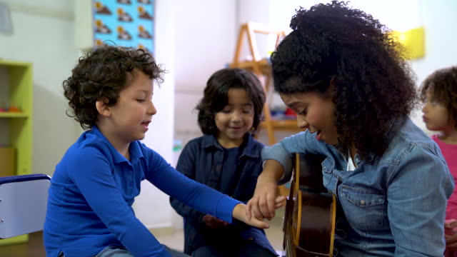 Cheerful Latin American teacher teaching a schoolboy how to play the guitar while everyone else pays attention