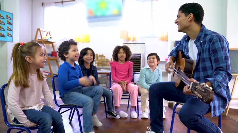 Male music teacher playing the guitar while all the class pays attention smiling and having fun