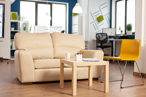 Modern business relax zone interior with confortable couch and orange chair. Rest area in company center with small sofa, shot of empty room with modern furniture, white shelves and blue wall.