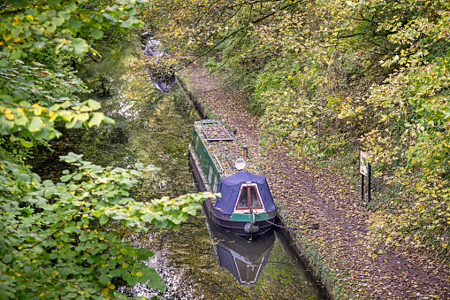 Tring, Hertfordshire, England - November 3nd 2023:  Houseboat moored in a narrow channel going through a small autumn forest