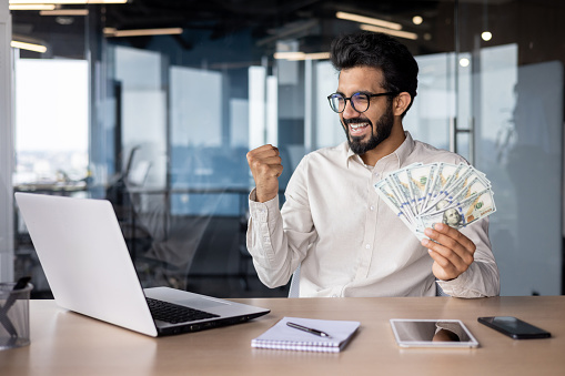 Young indian man working in office at laptop, holding money cash, happy with success, showing victory gesture with hand.