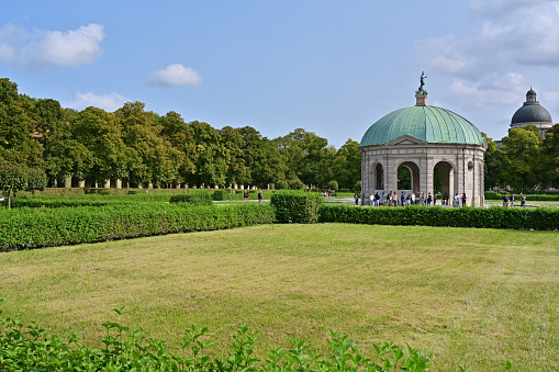 Photo showing a neatly mown and very green garden lawn, edged by a mature border containing a mixture of shrubs / evergreen plants and herbaceous flowers.  They include a number of plants with yellow flowers, such as daylilies (hemerocallis), cotton lavender (santolina) and lilies, as well as clipped topiary yew trees and box hedging.