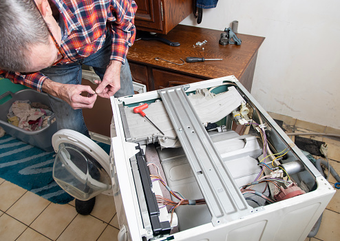 middle-aged master in a checkered suit unscrews the lid of a washing machine with a screwdriver for repair, repairs washing machines and household appliances at home, high quality photo