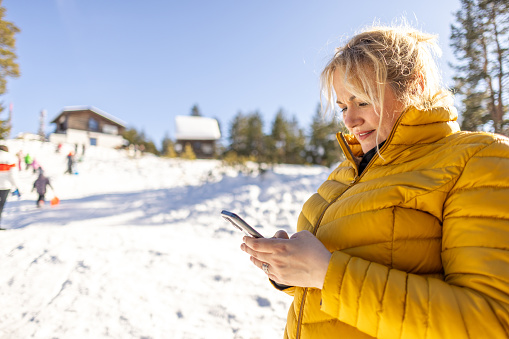 Side-view of a Caucasian mid-adult woman using a mobile phone, during her winter vacation on the mountain