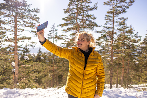 Caucasian mid-adult woman taking selfie with mobile phone, at the forest, during her winter vacation