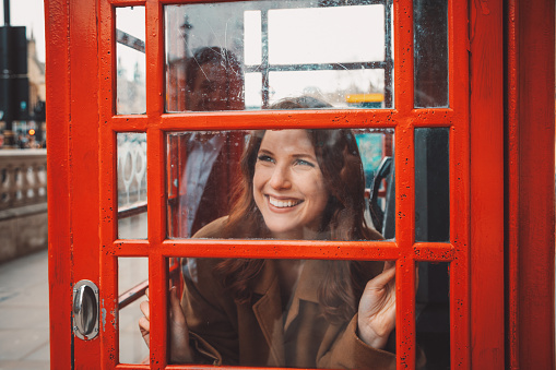 In the enchanting streets of London, a joyful and beautiful young tourist woman stands inside the iconic red telephone booth, gazing out onto the bustling street. Her eyes light up with happiness as she catches sight of her beloved boyfriend through the reflective glass of the booth. A warm smile plays on her lips as she witnesses a tender moment, sharing a connection that transcends the barriers of the telephone booth. The vibrant atmosphere of London's streets seems to fade into the background, leaving only the magic of their shared laughter and the timeless beauty of love.