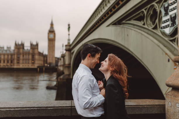 happy and young tourist couple kissing and enjoying a romantic getaway in iconic streets of london city against big ben, england, united kingdom - romantic getaway stock-fotos und bilder