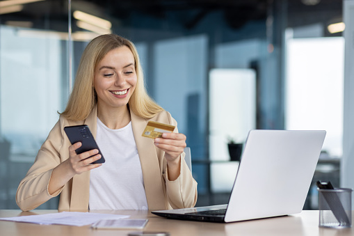 Young joyful woman at workplace inside office holds phone , bank credit debit card in hands, businesswoman chooses gifts in online store, book service and transfer funds using app on smartphone.