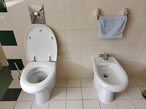 White toilet and bidet in the toilet. Above them is a towel and a water drain button. Ceramic tiles on the walls. Example of bathroom decor and interior