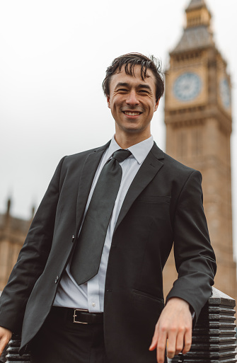 Capturing the essence of a modern businessman's journey, this image features a young and handsome man in a sharp black suit and tie, flashing a confident smile against the iconic backdrop of Big Ben or Elizabeth Tower in London, UK. 
Business Traveler, Young Man, London Trip, Stylish Businessman, Big Ben, Cityscape, Iconic Landmarks, Urban Elegance, Professional Style, Business Fashion, Modern Entrepreneur