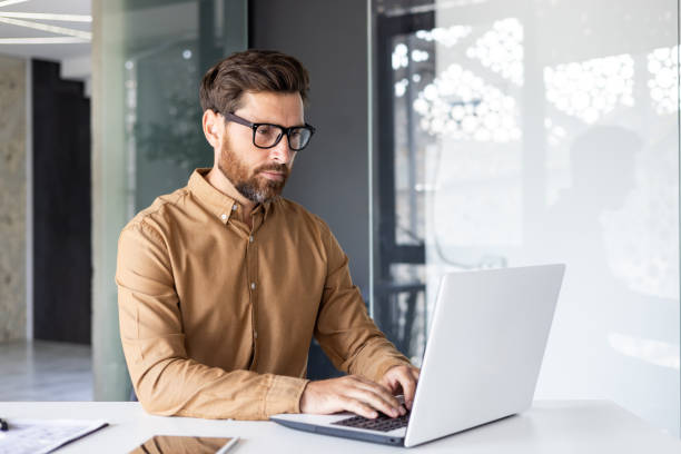 istock Serious concentrated thinking businessman working inside office with laptop, mature enlightened man in shirt and glasses at workplace typing on keyboard, programmer developer 1858004740