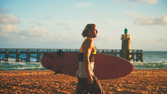 Handsome man surfer carrying surfing board on beach during sunset