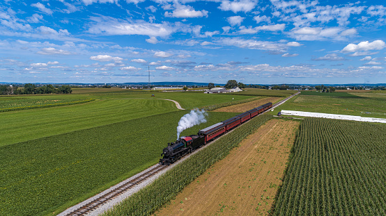 A Drone Frontal View of a Restored Steam Passenger Train, Traveling Thru Green Corn Fields, Blowing Smoke on a Fall Day